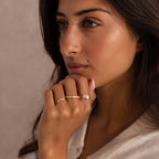 Woman with long dark hair wearing three gold rings, including one with a pear-shaped opal and rectangular clear gemstone, one with small pearls, and one with embedded clear stones.