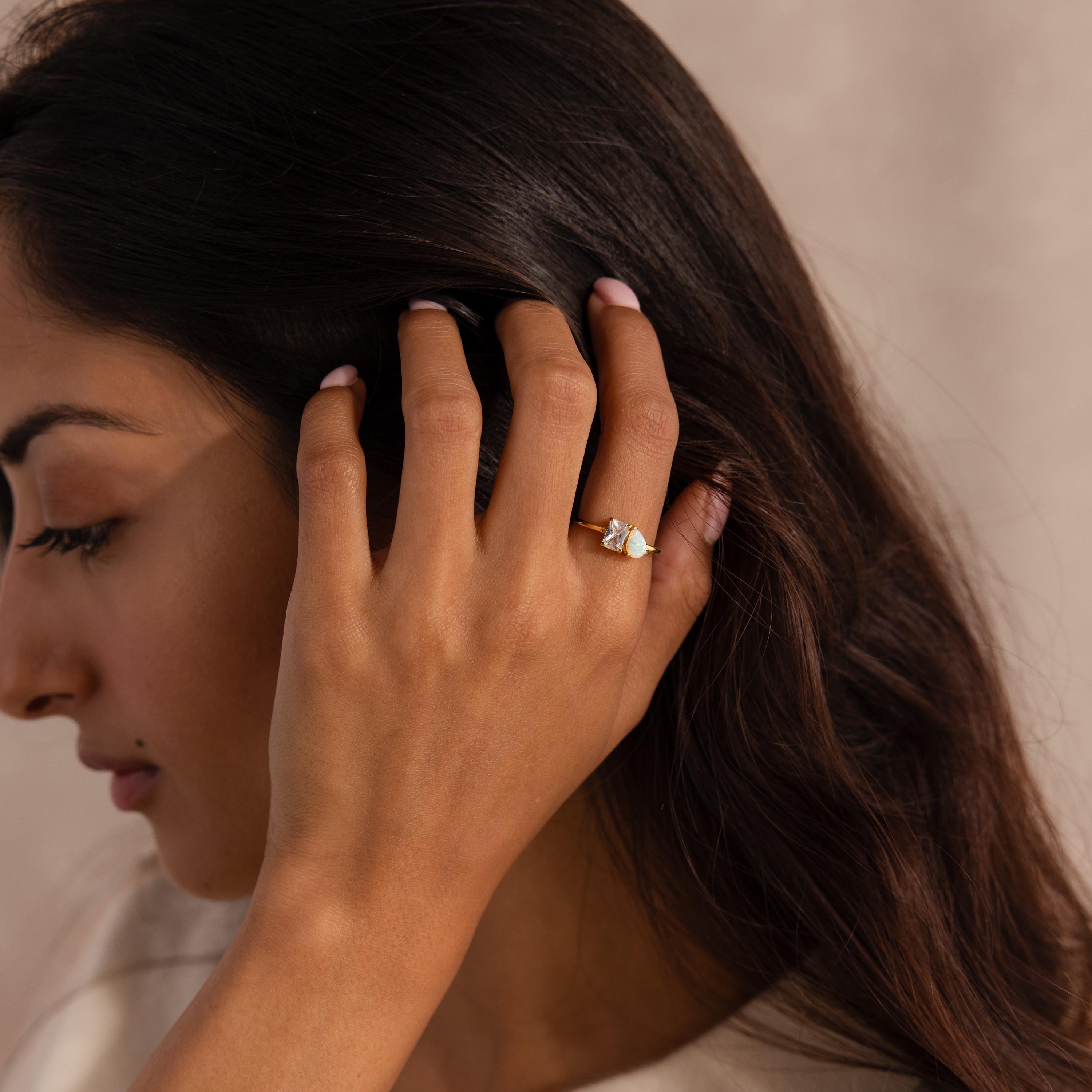 Woman with long dark hair tucking it behind her ear, wearing a gold ring with a rectangular clear gemstone and a pear-shaped opal.