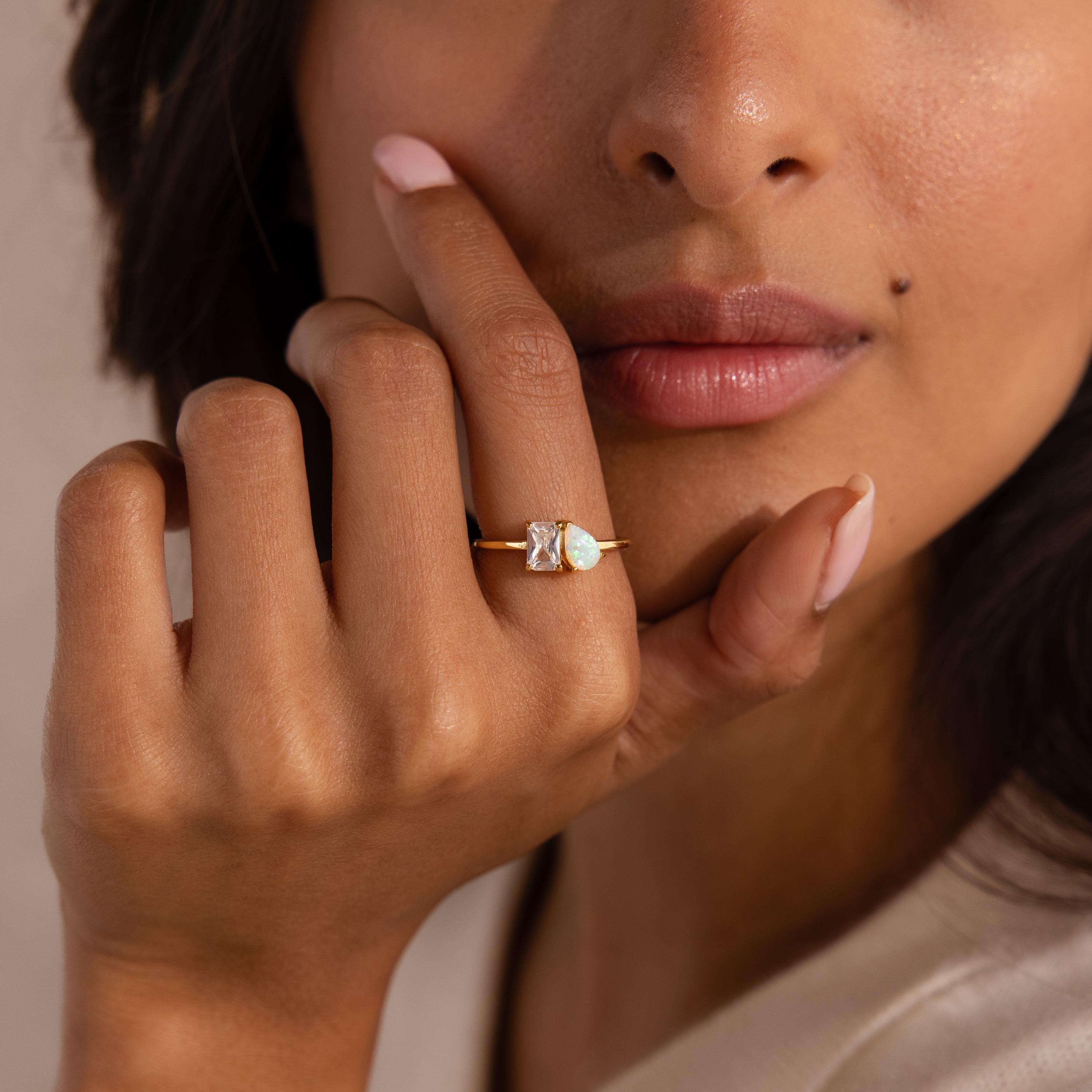 Close up of a woman’s hand near her lips, wearing a gold ring with two stones, a Baguette cut clear gemstone and a Pear Cut opal, set side by side on a thin band.