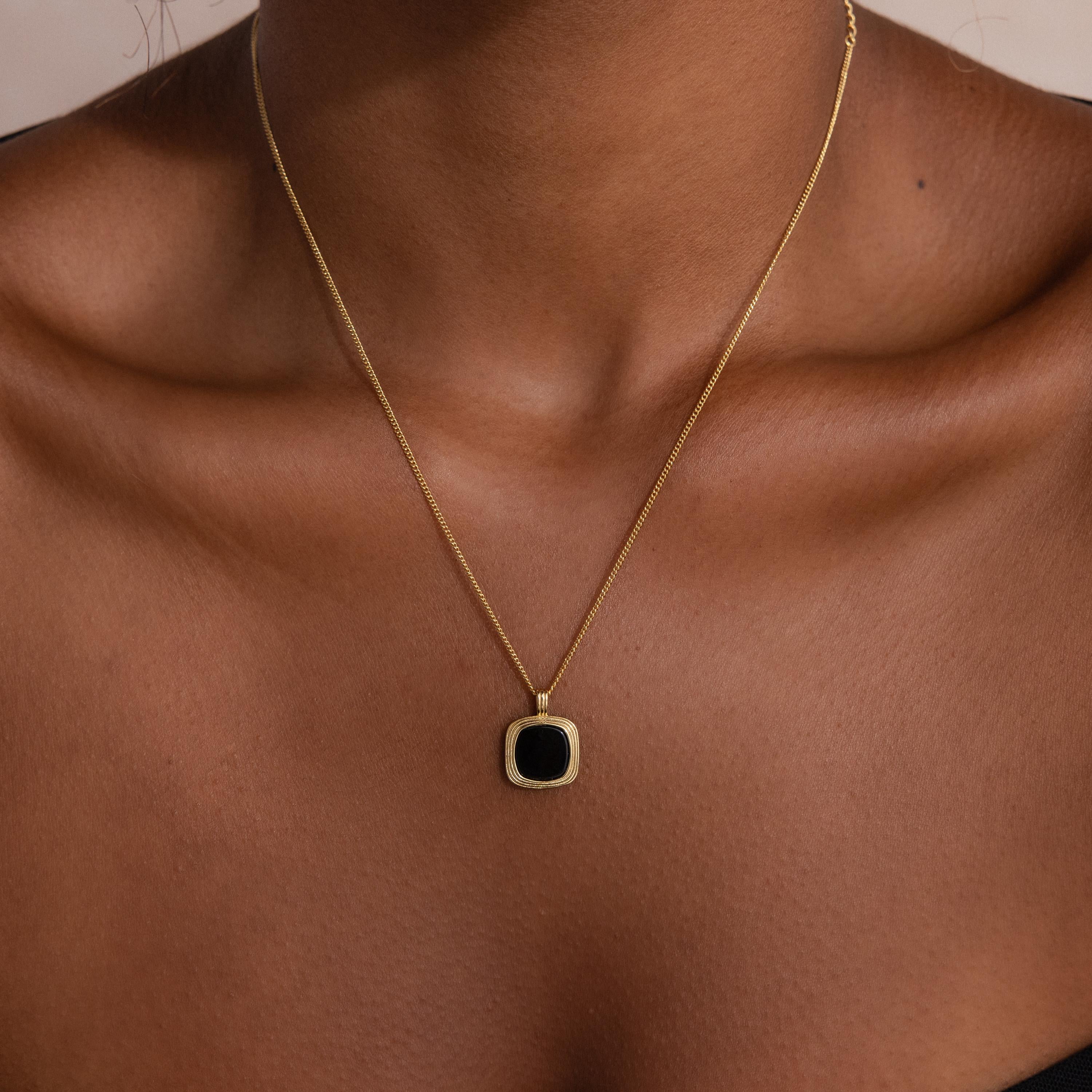 Close up of a woman's collarbone wearing a delicate gold chain necklace with a small square black onyx pendant framed in a textured gold bezel setting.