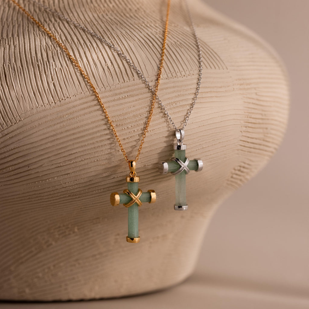 Two jade cross pendant necklaces, one with gold end caps and chain, and the other with silver end caps and chain, both featuring an X shaped metal accent at the center, displayed on a textured beige surface.