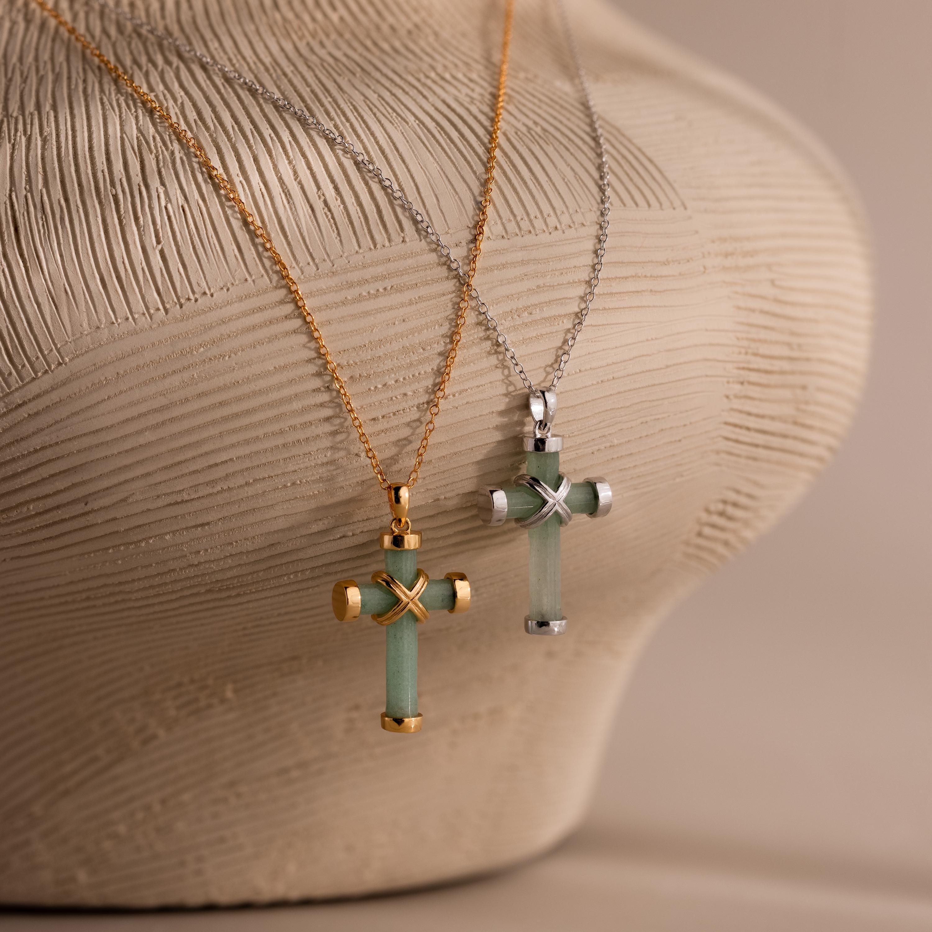 Two jade cross pendant necklaces, one with gold end caps and chain, and the other with silver end caps and chain, both featuring an X shaped metal accent at the center, displayed on a textured beige surface.