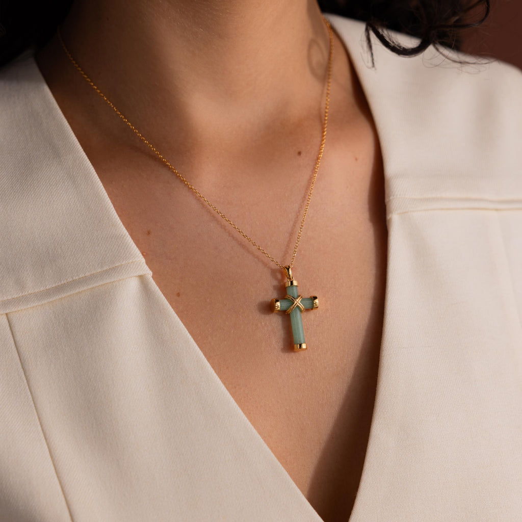 Gold chain necklace with a green jade cross pendant featuring polished gold end caps and a gold X shaped accent, worn with an ivory V neck blouse.