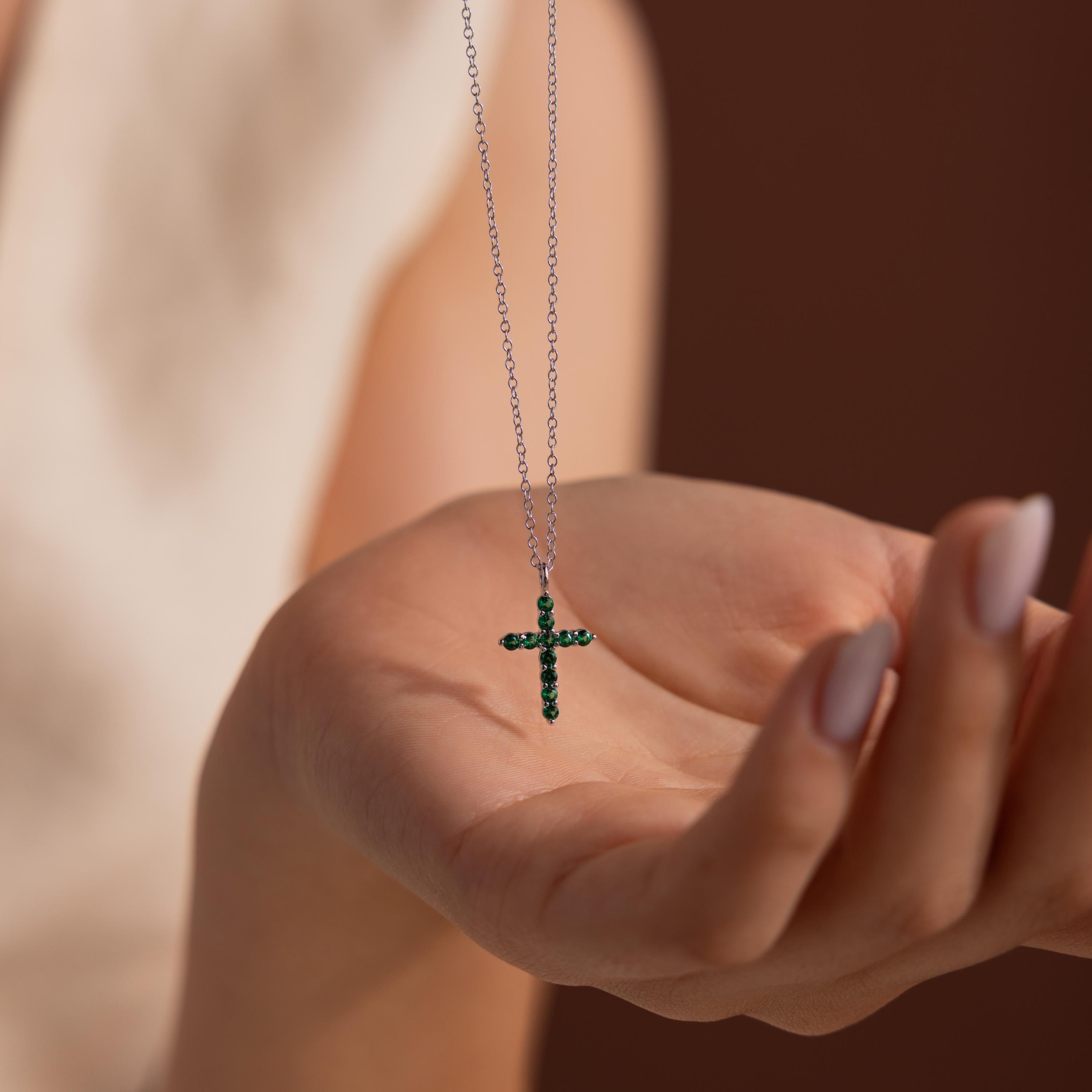 Silver chain necklace with a cross pendant set with small round green gemstones, displayed resting in the palm of a hand with a blurred neutral background.