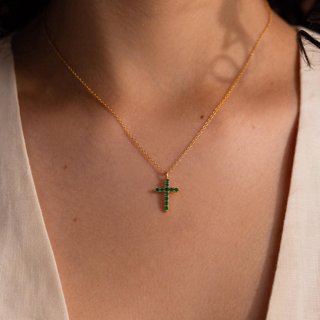Gold chain necklace featuring a small cross pendant adorned with green gemstones, worn against bare skin with a cream V neck top.