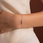 Silver chain bracelet worn on the wrist, featuring a delicate cross shaped charm adorned with round white gemstones.