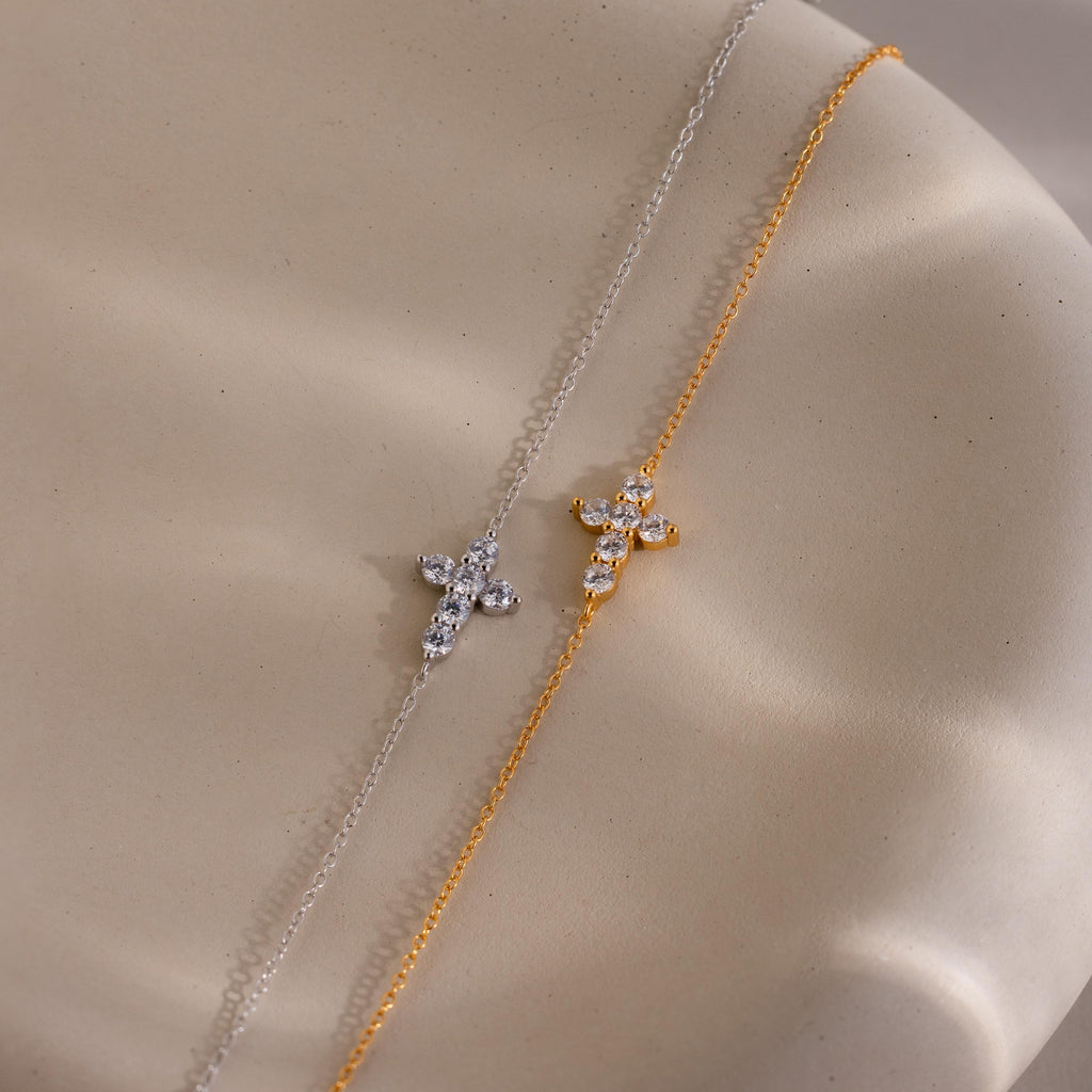 Two delicate chain bracelets displayed side by side, one in silver and one in gold, each featuring a small cross shaped charm adorned with round cut clear gemstones.