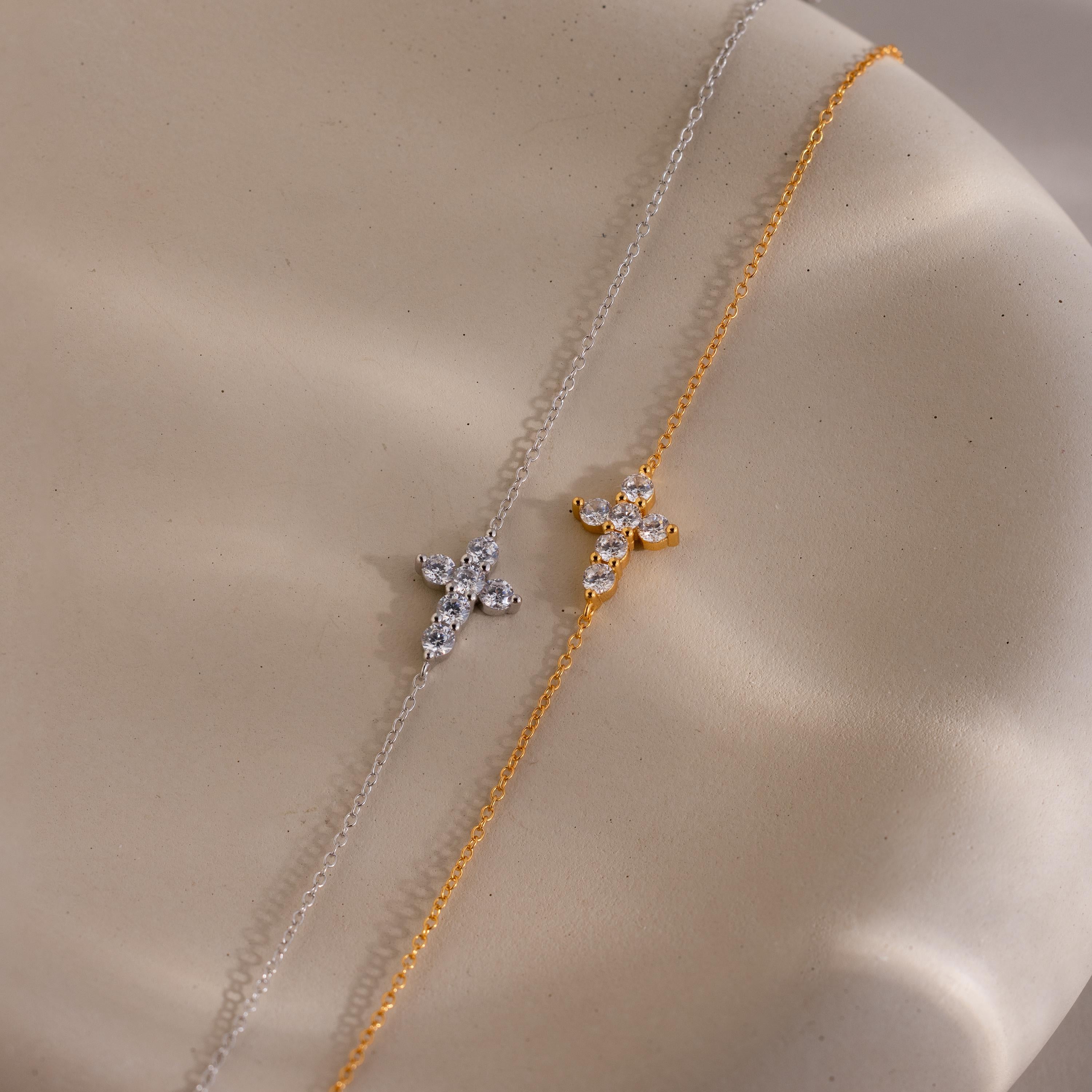 Two delicate chain bracelets displayed side by side, one in silver and one in gold, each featuring a small cross shaped charm adorned with round cut clear gemstones.