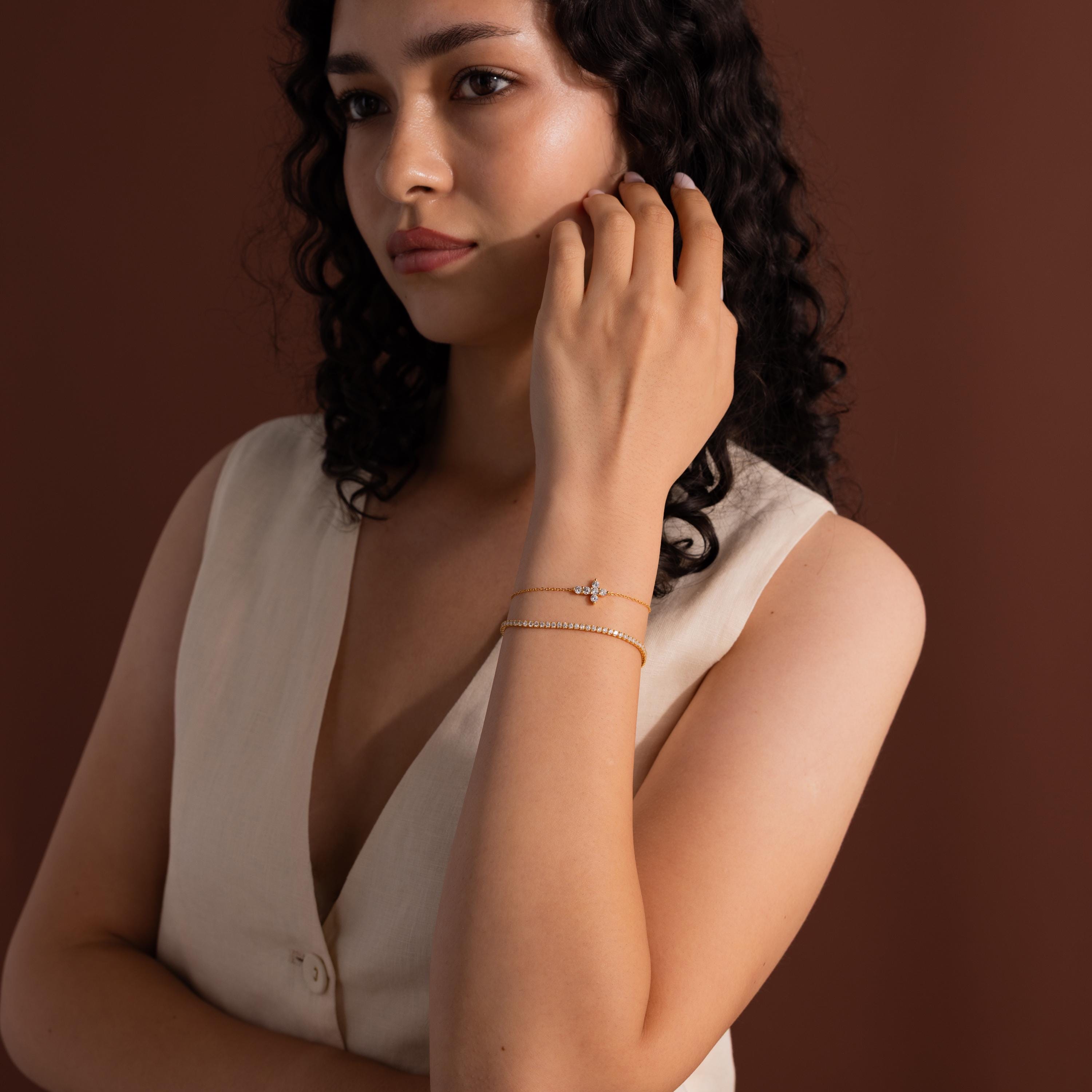 Woman with curly hair wears a cream sleeveless top and dainty jewelry, including a gold chain and the Diamond Cross Bracelet, as she poses against a brown background.