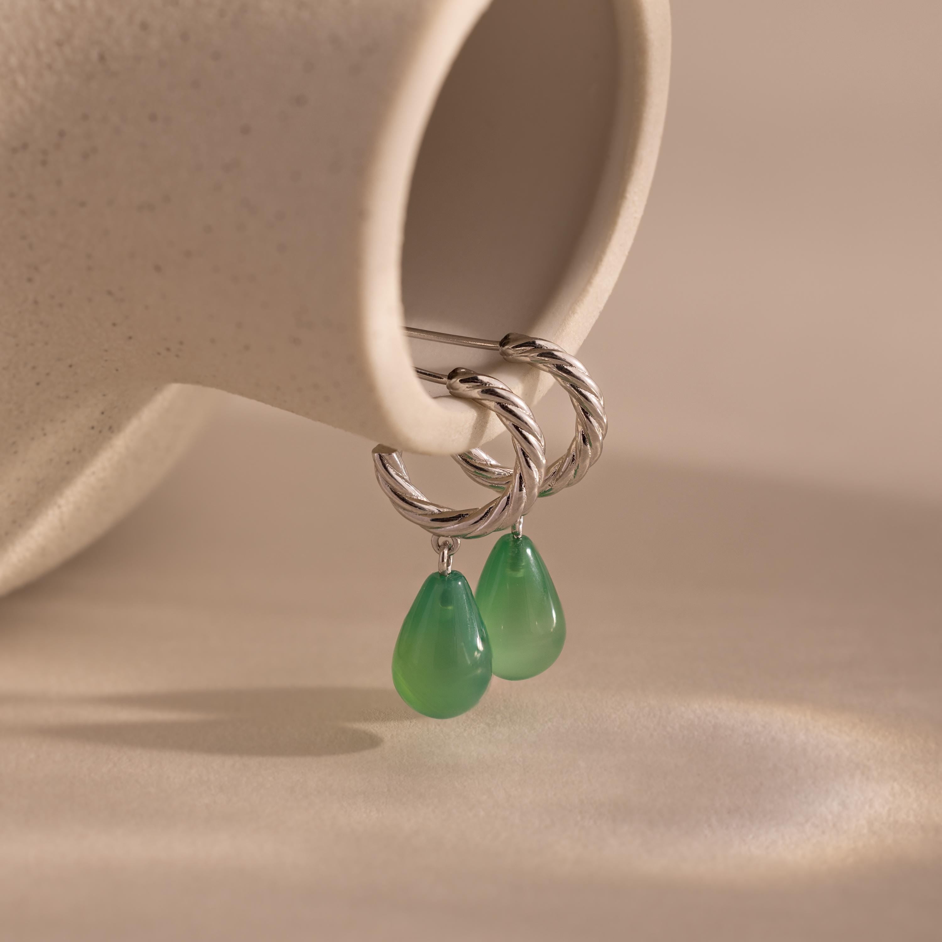 Pair of twisted silver hoop earrings with green jade teardrop charms displayed on a smooth beige background.