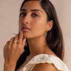 Woman wearing twisted gold hoop earrings with green jade drops, paired with gold rings and an ivory scalloped top.