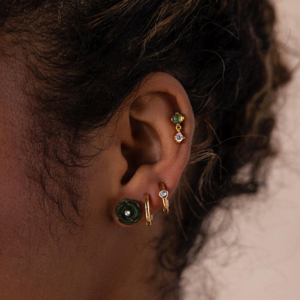 Close-up of a woman’s ear adorned with multiple earrings, highlighting a green carved jade rose stud with a sparkling center stone.
