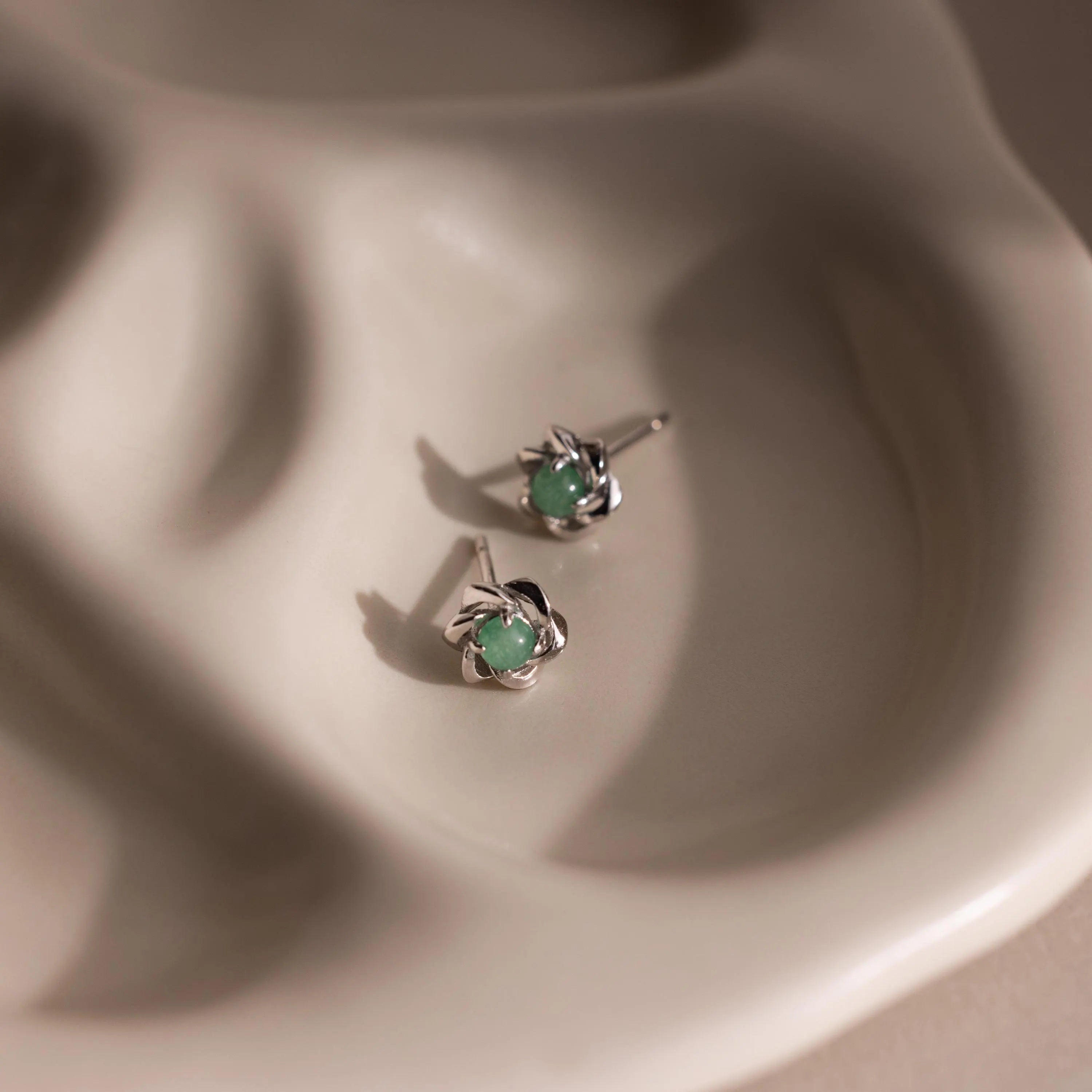 Flay-lay of silver flower-shaped stud earrings with a round green jade gemstone center.