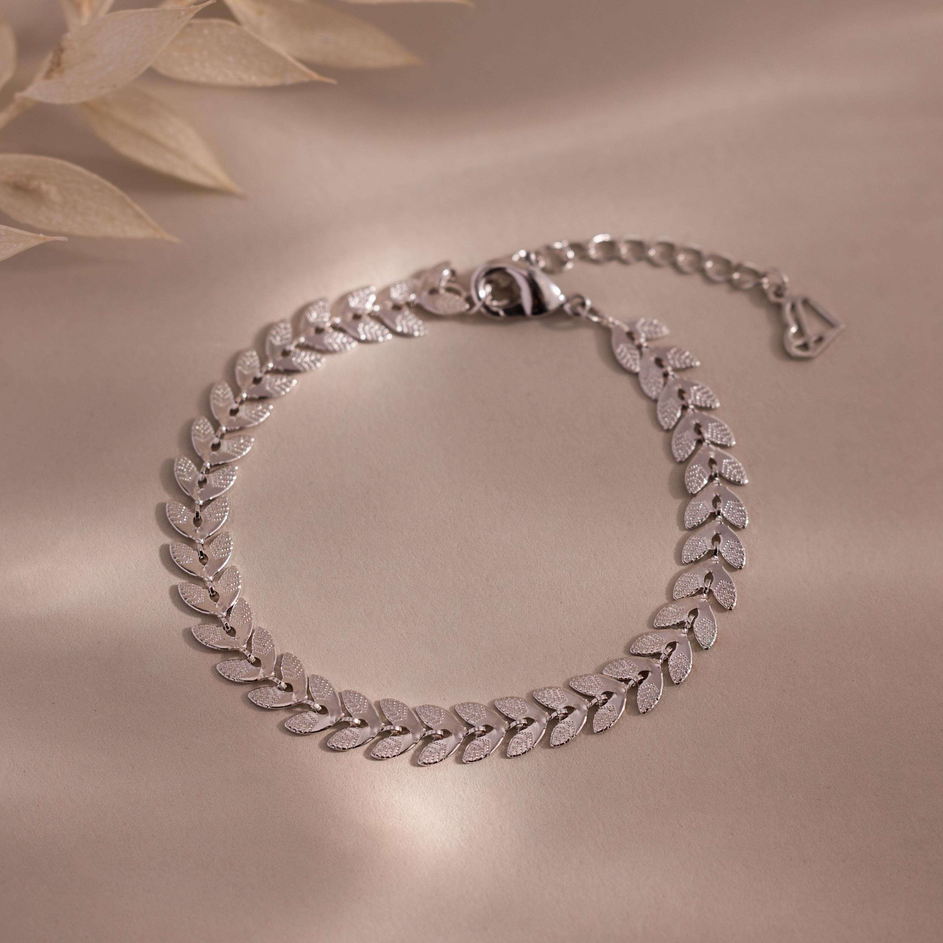 Flat-lay of a white gold leaf link chain bracelet with textured leaf-shaped links displayed on a neutral surface.