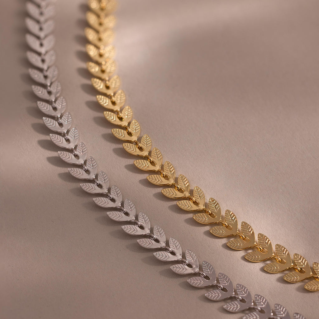 Close-up of two leaf link chain bracelets in gold and white gold, each with textured leaf-shaped links, displayed on a neutral surface.