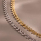 Close-up of two leaf link chain bracelets in gold and white gold, each with textured leaf-shaped links, displayed on a neutral surface.