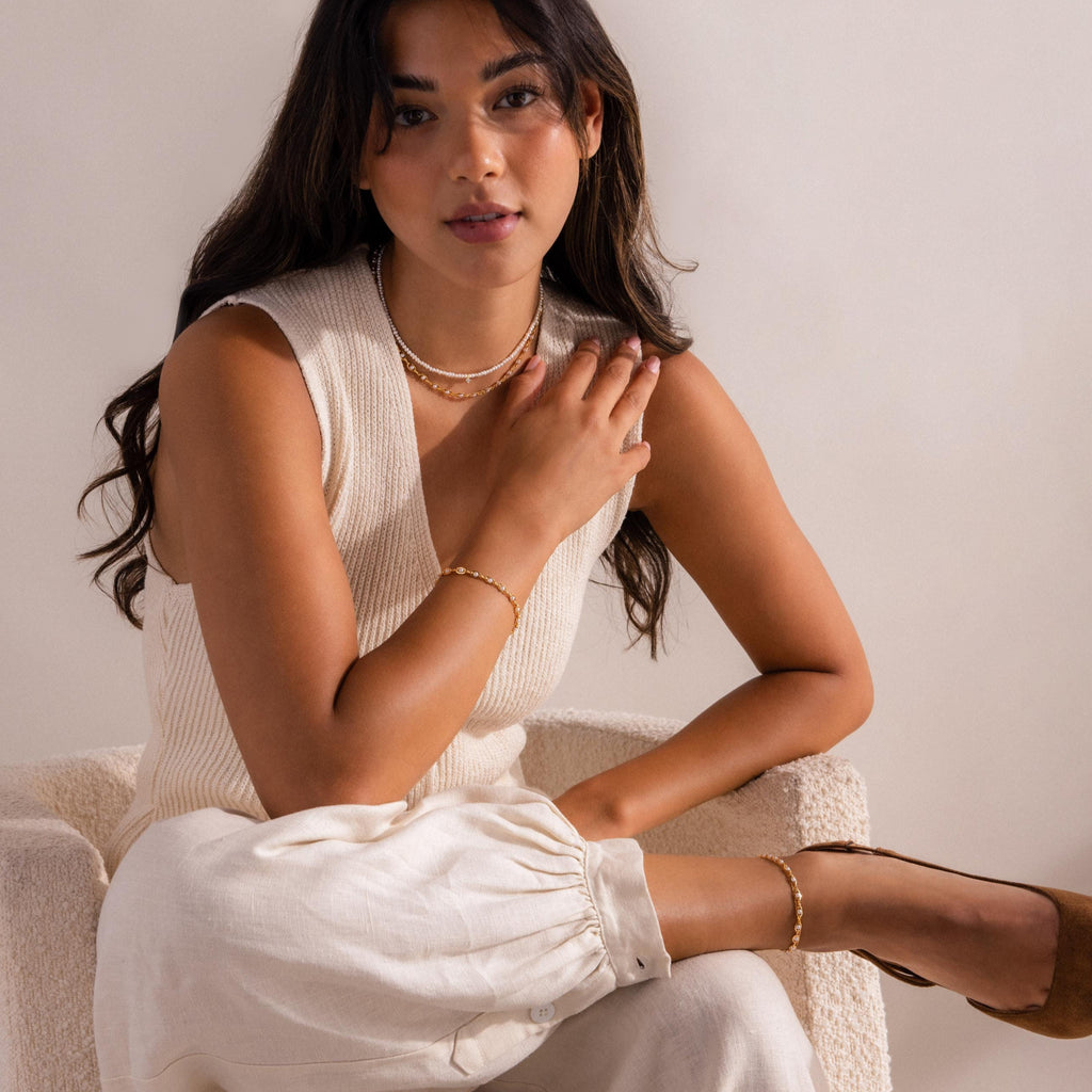 Woman wearing layered gold and pearl jewelry including a necklace, bracelet, and anklet, styled with a sleeveless cream knit top, linen pants, and brown suede heels while seated on a textured chair.