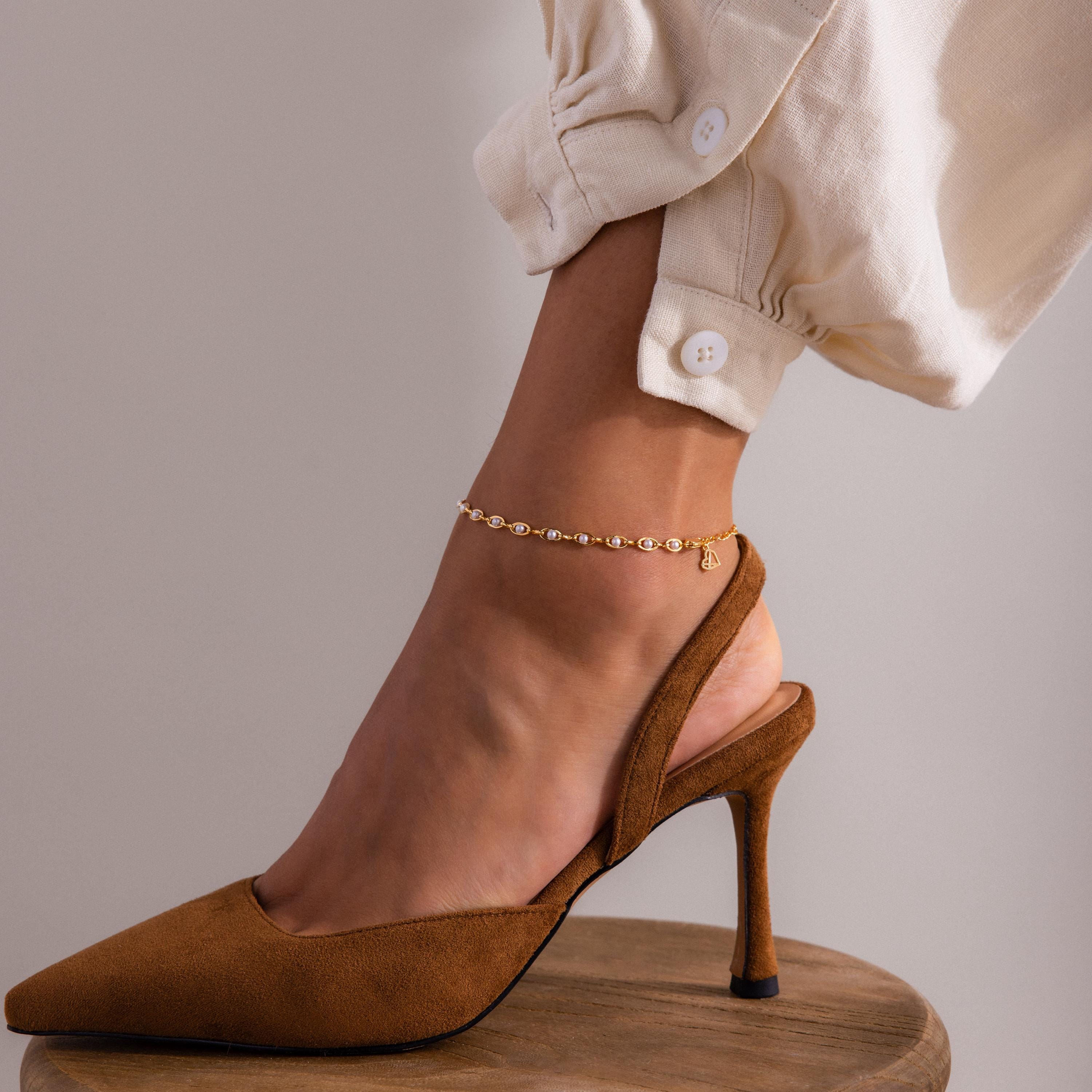 Gold anklet with small pearls and delicate chain links, worn around the ankle and styled with tan suede high heels and cream cuffed pants.