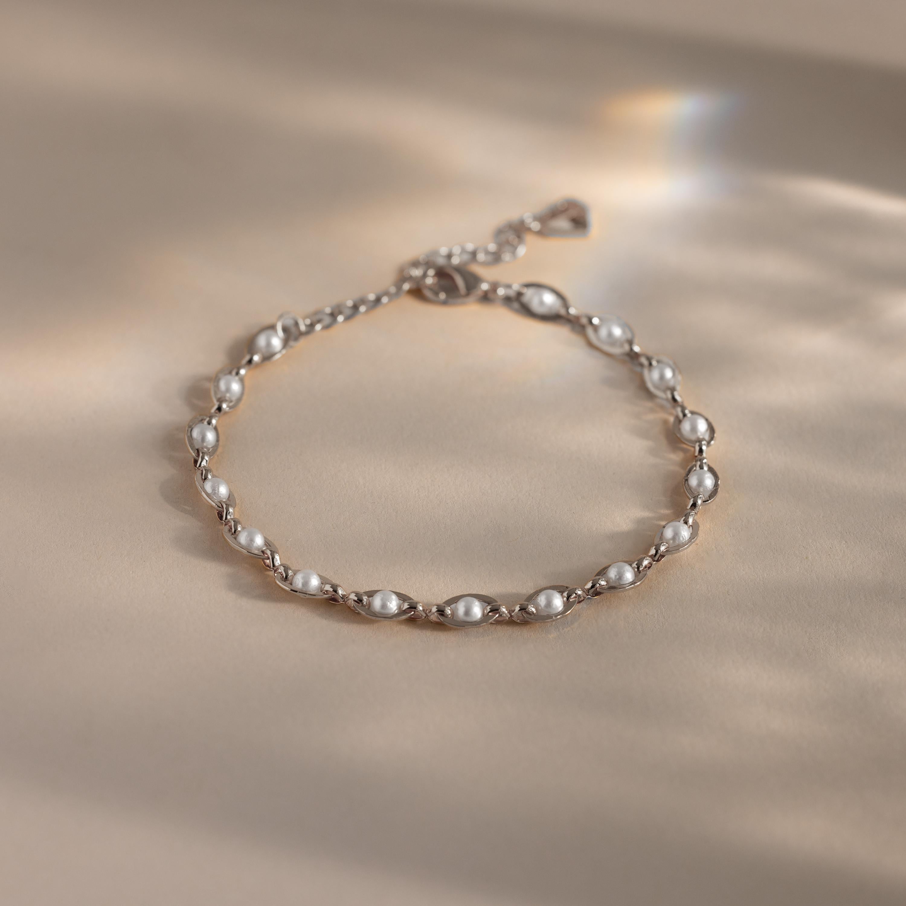 Silver chain bracelet with evenly spaced white pearls, featuring an adjustable clasp and heart-shaped detail, displayed on a neutral surface with soft light reflections.