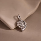 Silver shell-inspired charm pendant with a textured circular design and gemstone accents, displayed on a soft neutral fabric background.