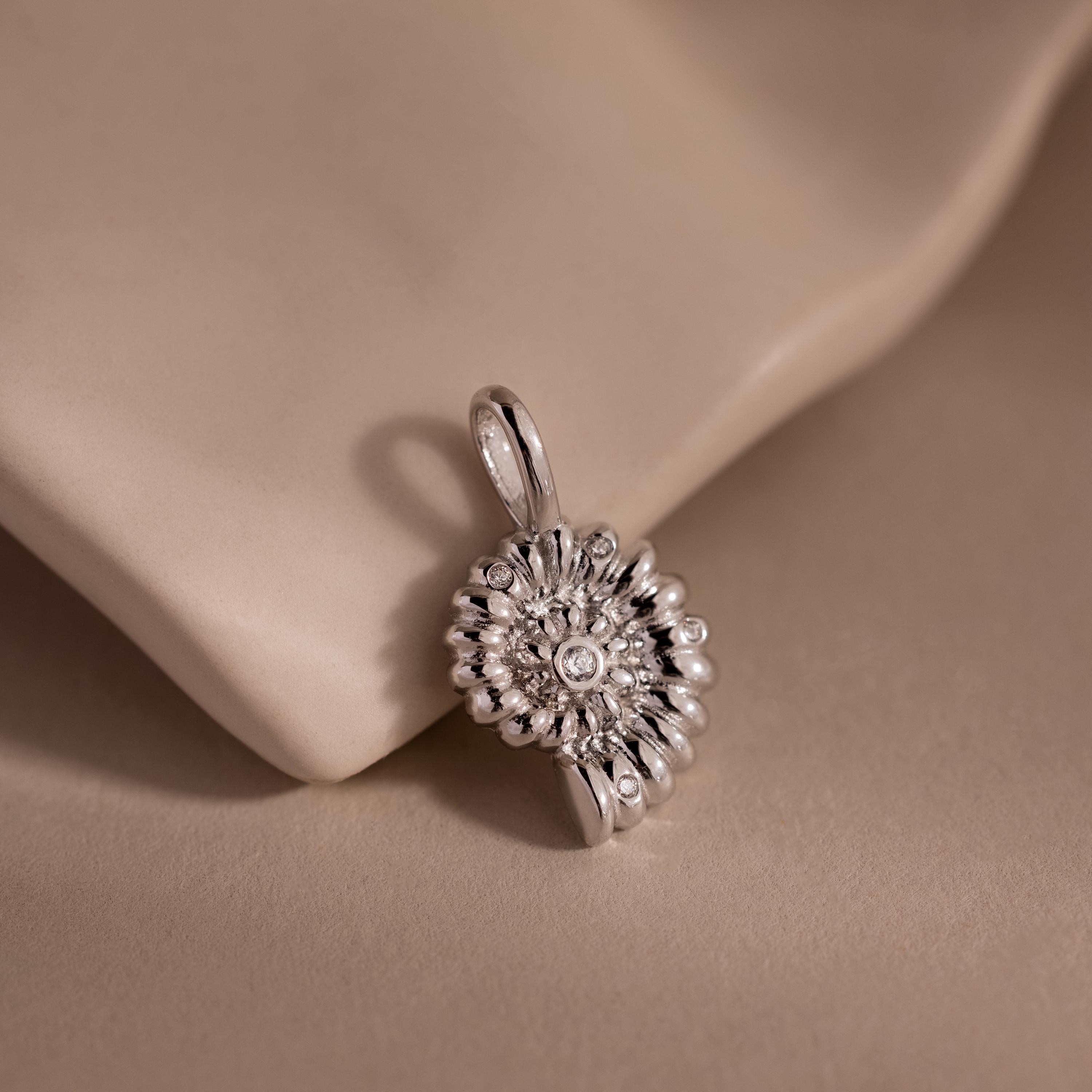 Silver shell-inspired charm pendant with a textured circular design and gemstone accents, displayed on a soft neutral fabric background.