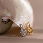 Gold and silver shell inspired charm pendants with textured circular designs and gemstone accents, displayed side by side against a large iridescent seashell.