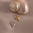 Two Shark Tooth Charm pendants—one gold talisman, one silver—rest by a seashell on a neutral backdrop. These ocean-inspired beauties are stylish jewelry pieces to complete any look.