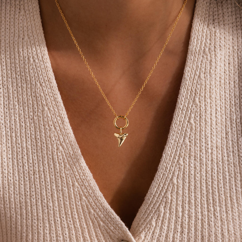 Gold shark tooth charm pendant on a delicate chain necklace, styled with a cream knit V-neck top.