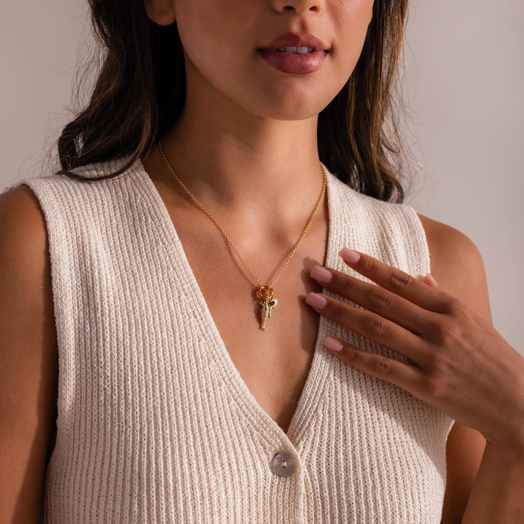 Gold chain necklace with layered charms including a shark tooth and fish pendant, styled with a sleeveless cream knit top.