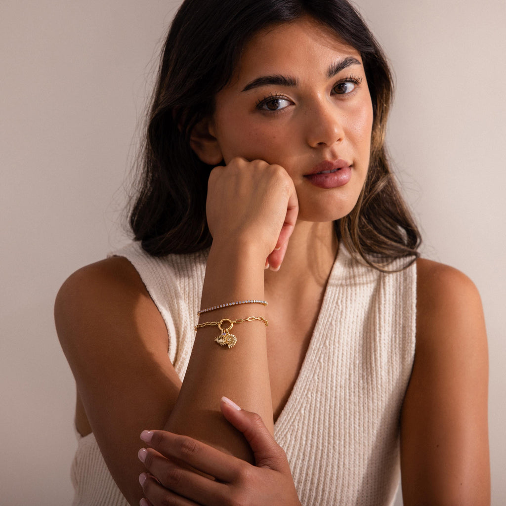 Woman wearing a gold charm bracelet with a seashell pendant, paired with a delicate beaded bracelet, styled with a sleeveless cream knit top.