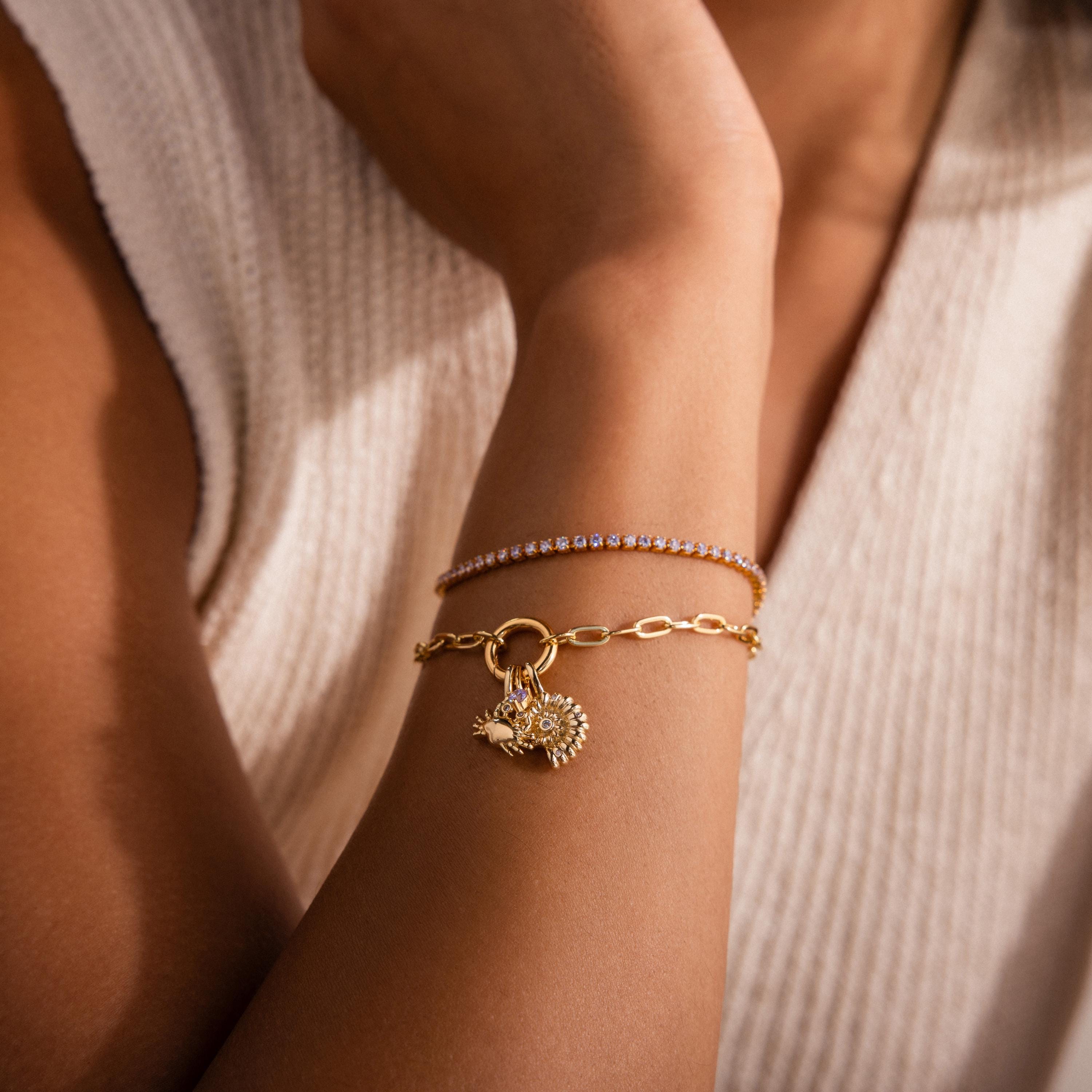 Woman wearing a gold charm bracelet with crab and seashell pendants, stacked with a delicate gemstone tennis bracelet, styled with a cream knit top.