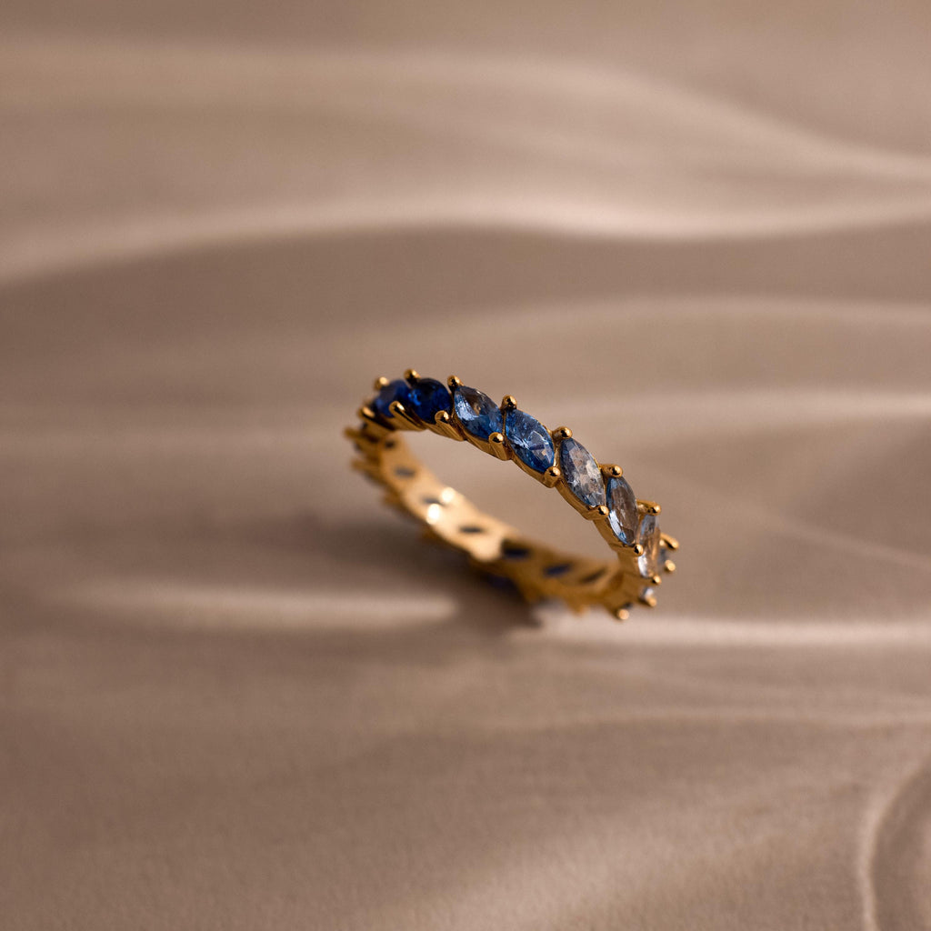 Single gold eternity ring displayed upright on a neutral surface, featuring marquise cut stones in a blue ombre gradient from deep navy to pale sky blue.