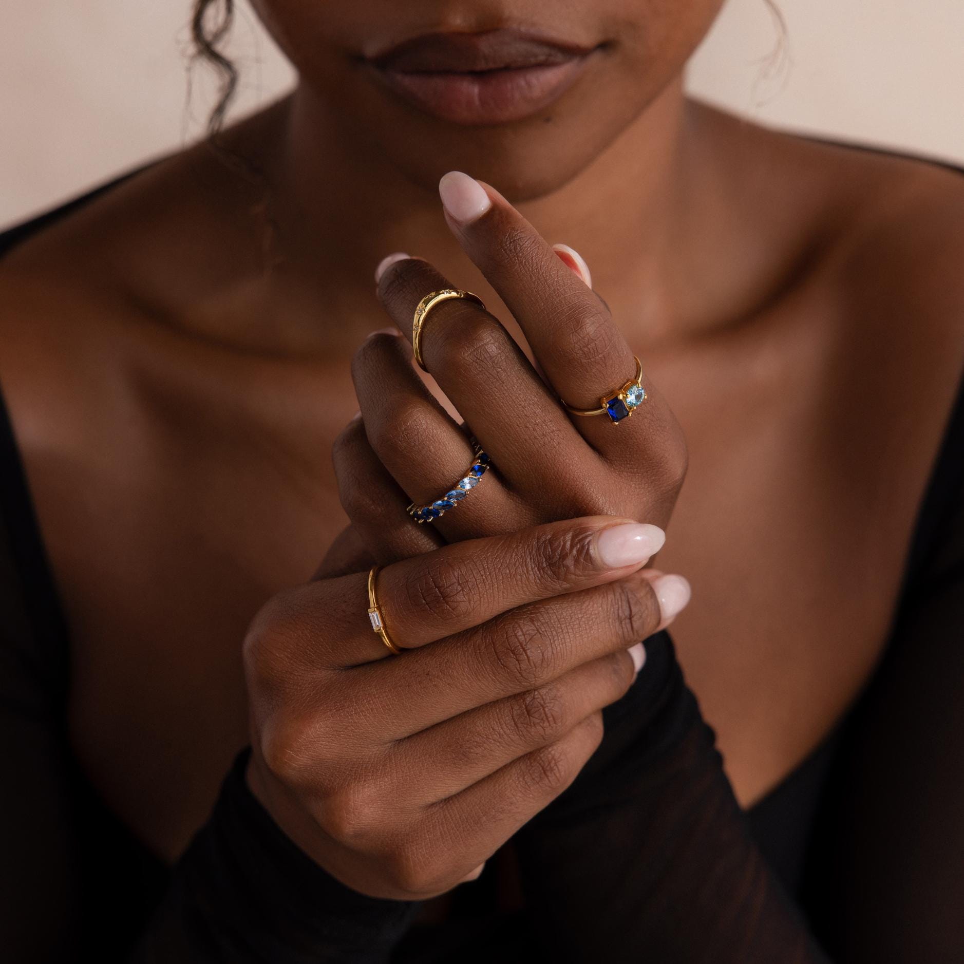 Model’s hands adorned with multiple gold rings, including a marquise cut blue ombré eternity band on the middle finger, a two stone blue and clear ring on the index, and minimal bands on other fingers.