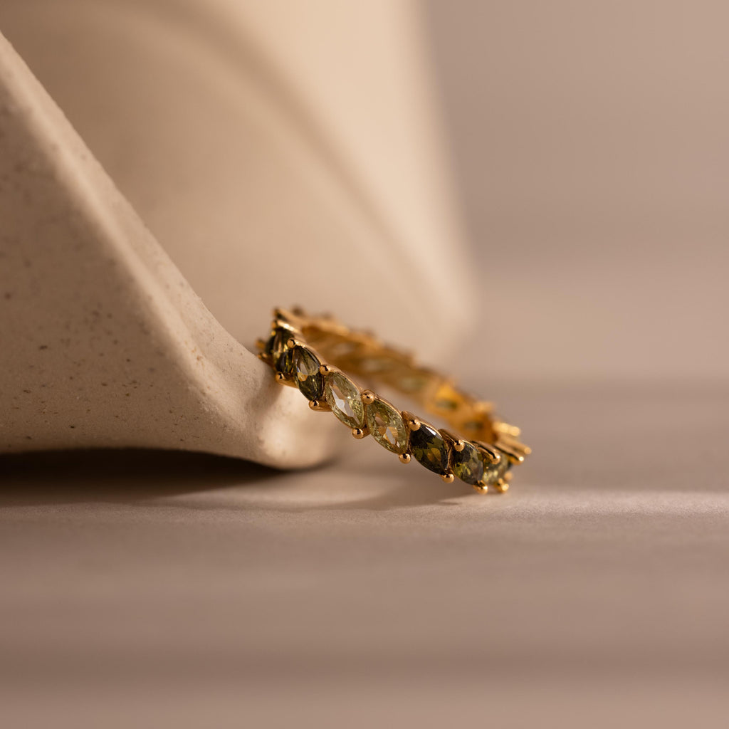 Gold eternity ring with marquise cut stones in ombre green tones, transitioning from pale lime to deep moss, rests against a sculptural ceramic form on a soft beige surface.