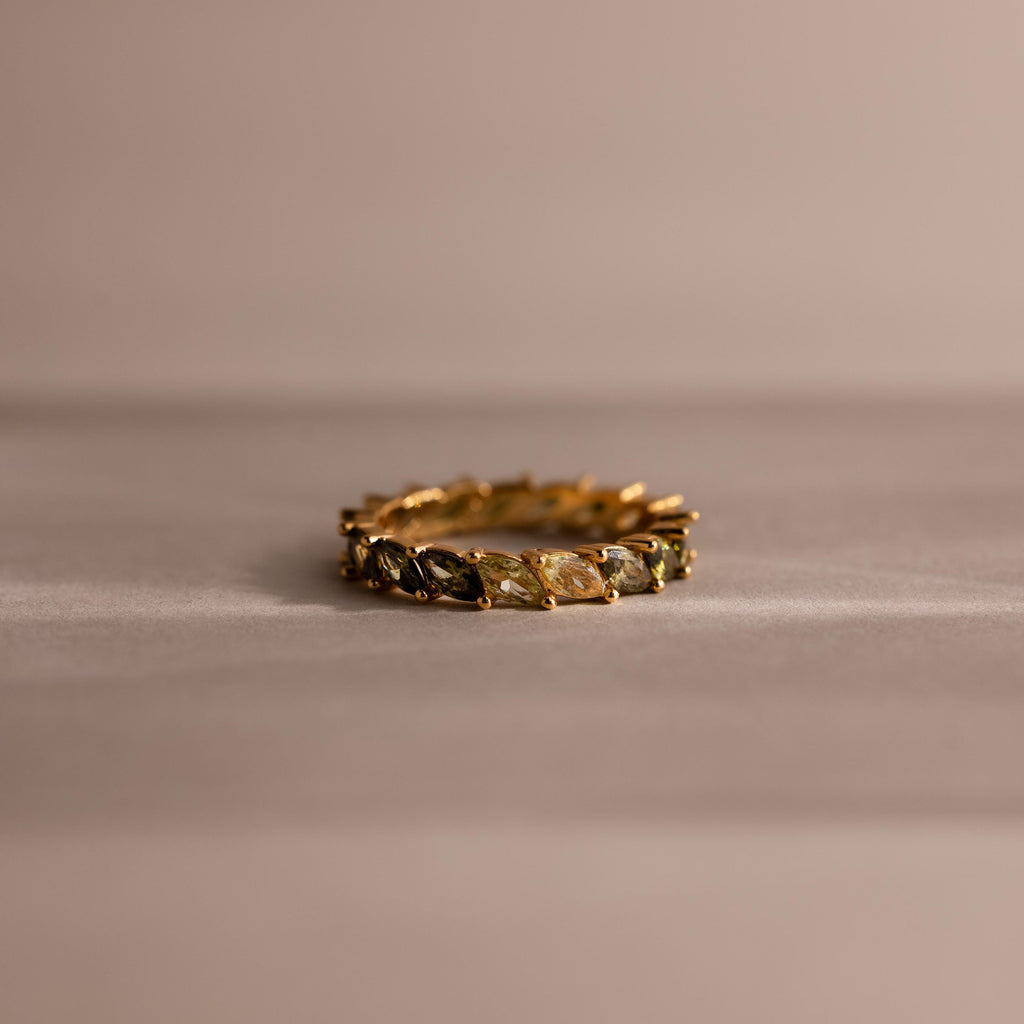 Marquise cut eternity ring with ombre green gemstones fading from deep olive to pale mint, set in a continuous gold band and displayed on a soft neutral surface.