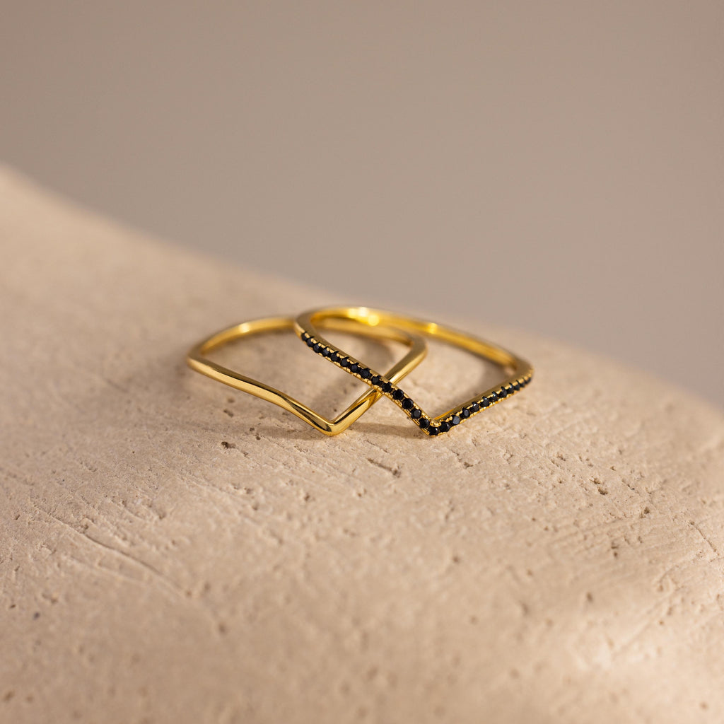 Two gold chevron rings displayed on a textured surface, one plain and one accented with pavé-set black stones.