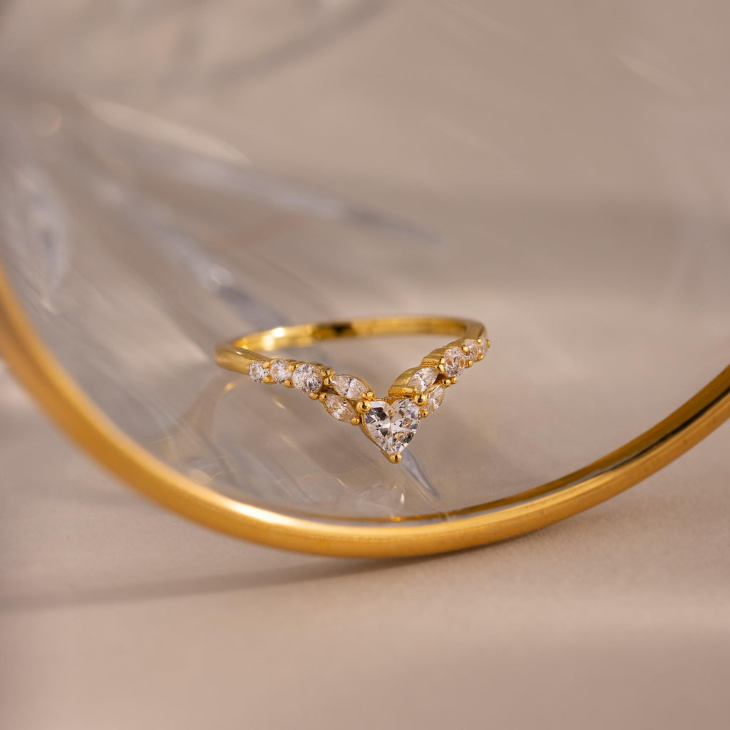 Gold heart curve cluster diamond ring with heart shaped center stone and round diamonds displayed against a reflective glass surface.
