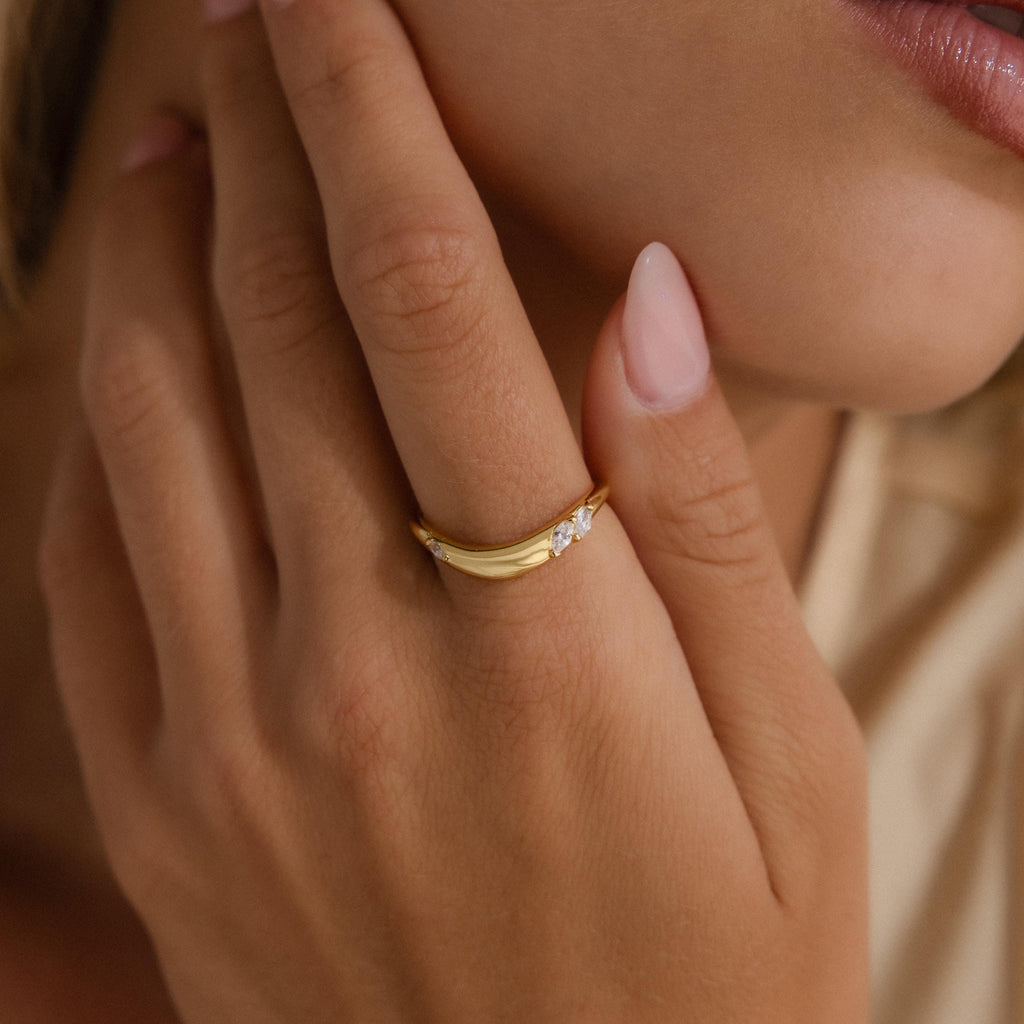 Minimalist gold curved band ring with three marquise cut clear gemstones, displayed on a textured stone surface.