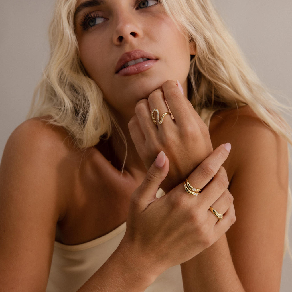 Woman wearing multiple gold rings, including a wavy statement band and a curved gold ring with gemstones, styled with a neutral sleeveless top for an elegant jewelry look.