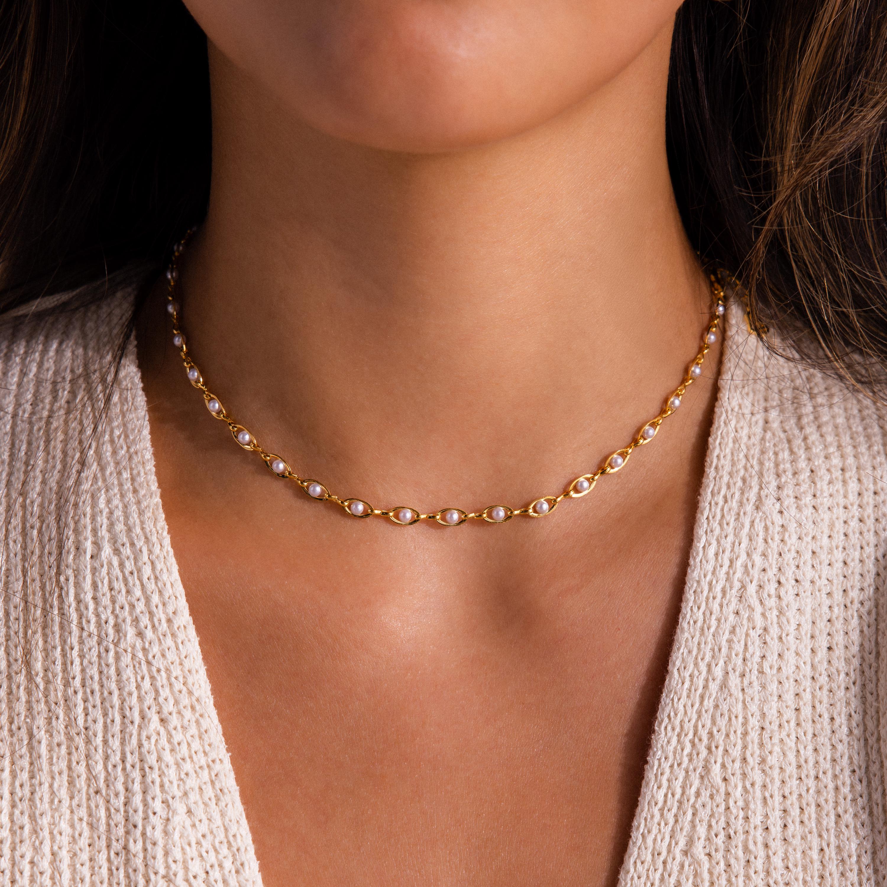 Gold choker necklace featuring evenly spaced white pearls, styled on a woman’s neckline with a textured cream knit top.