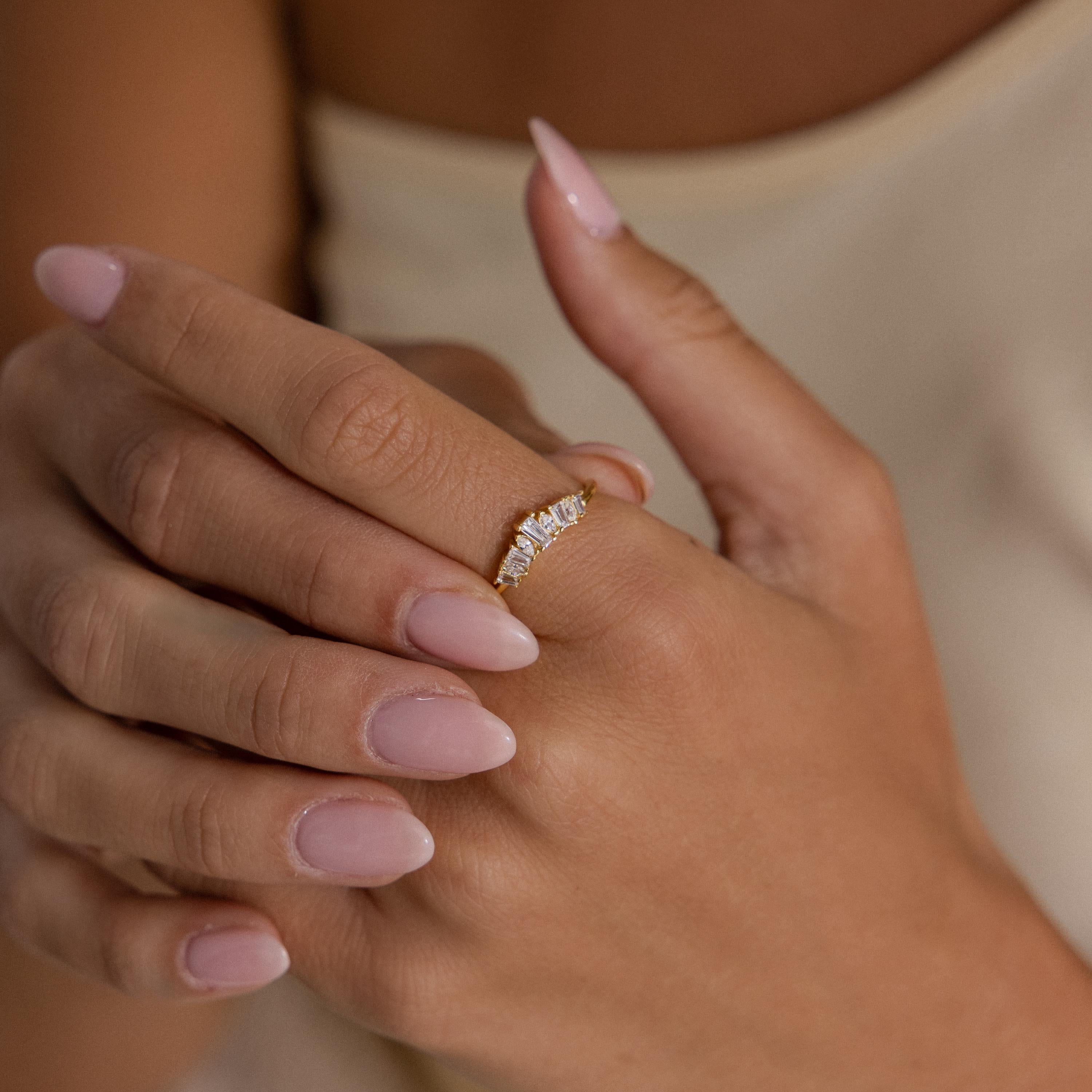 Close up of hands wearing a slim gold ring with a curved row of baguette and marquise cut clear stones, paired with soft pink manicured nails for an elegant and feminine look.