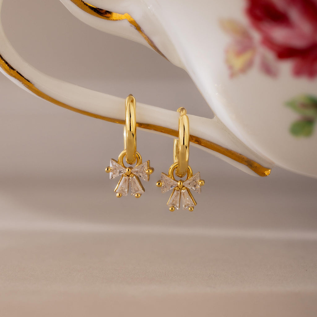Gold Starlight Bow Hoop earrings with dangling baguette cut clear stones arranged in a bow design, displayed hanging from a porcelain handle with floral detail.