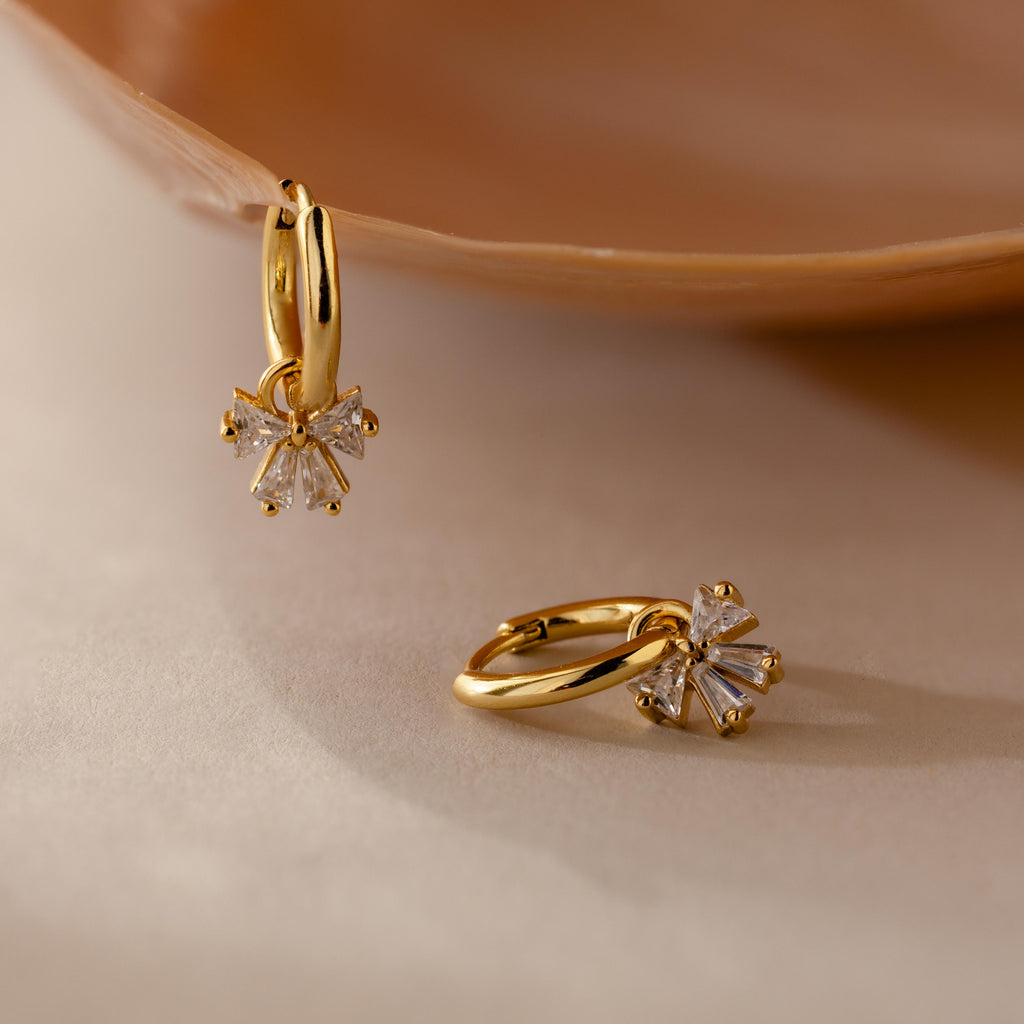 Gold Starlight Bow Hoop earrings with dangling baguette cut clear stones in a bow shaped design, displayed on a neutral surface for an elegant and modern look.