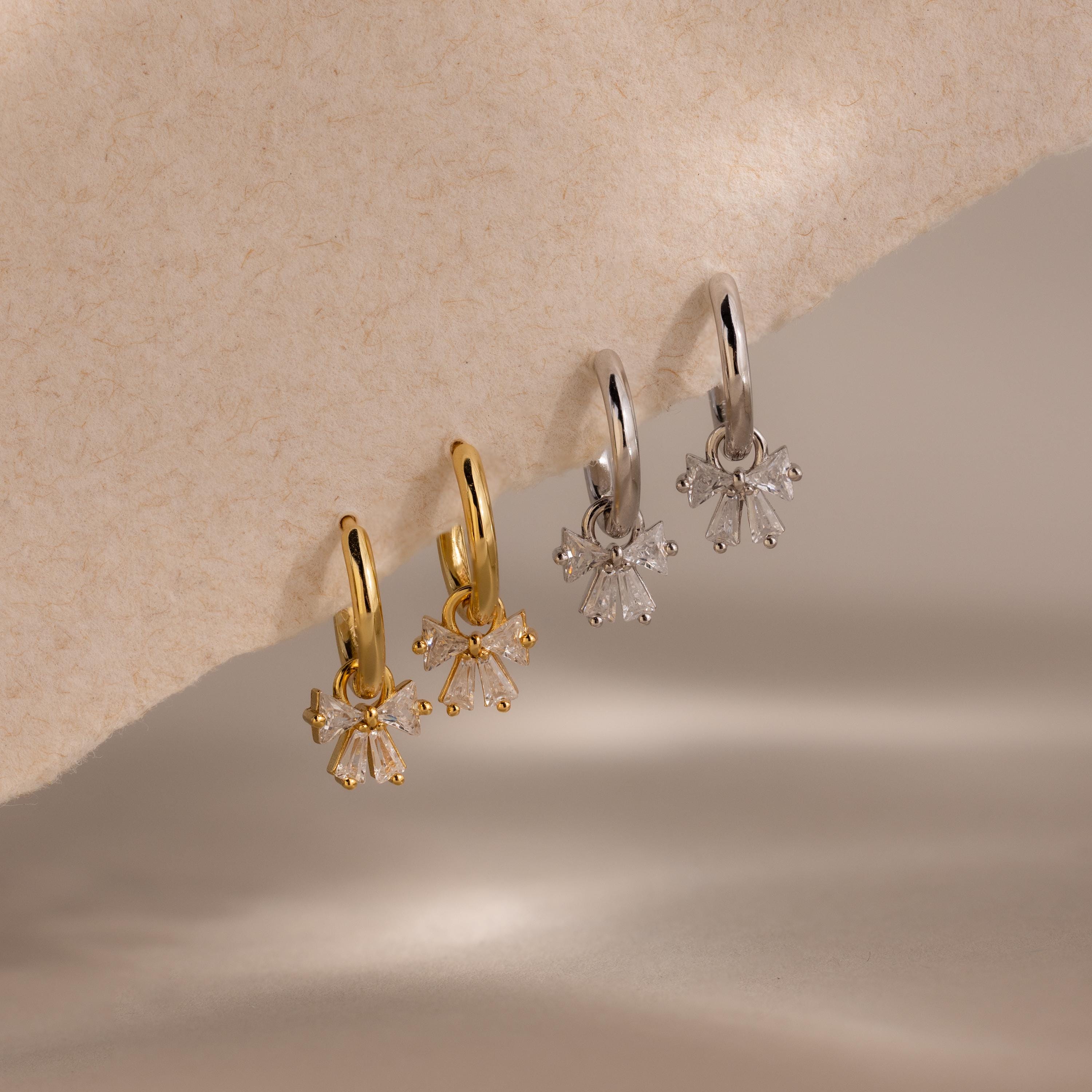 Two pairs of Diamond Bow Hoops—one gold, one silver—with clusters of clear gemstones hanging below; ideal for fans of bow earrings or anyone seeking sparkling elegance.