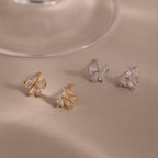 Two pairs of Diamond Bow Studs—one gold, one silver—feature butterfly-inspired designs reminiscent of diamond-cut bows, elegantly displayed on a beige surface.