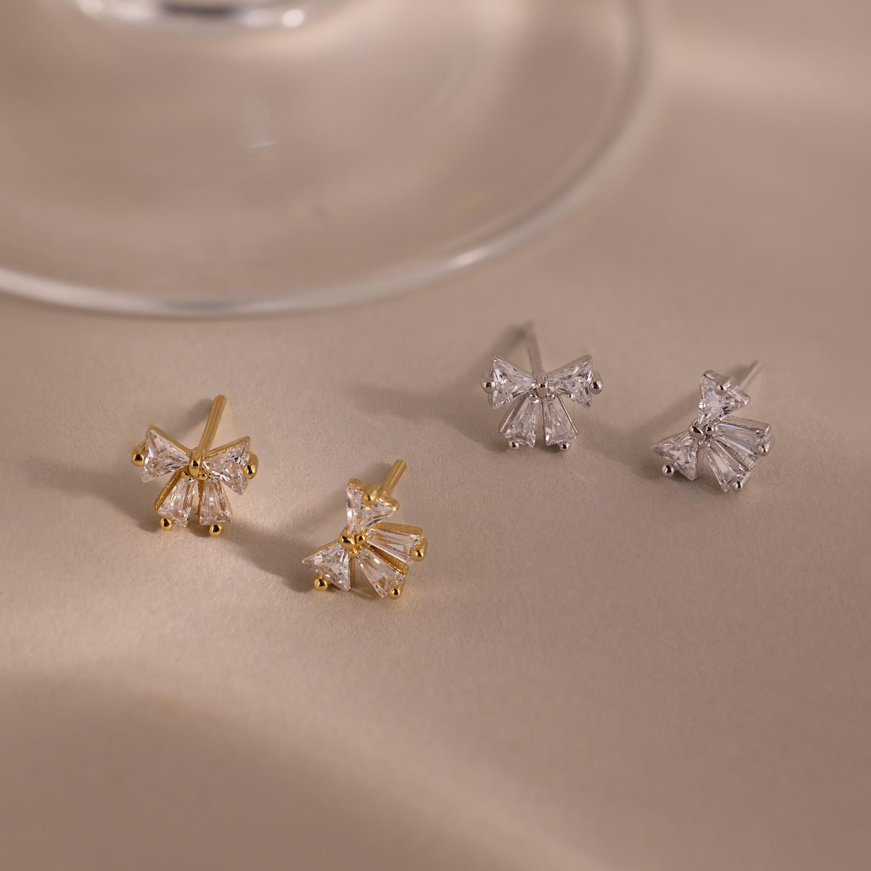 Two pairs of Diamond Bow Studs—one gold, one silver—feature butterfly-inspired designs reminiscent of diamond-cut bows, elegantly displayed on a beige surface.