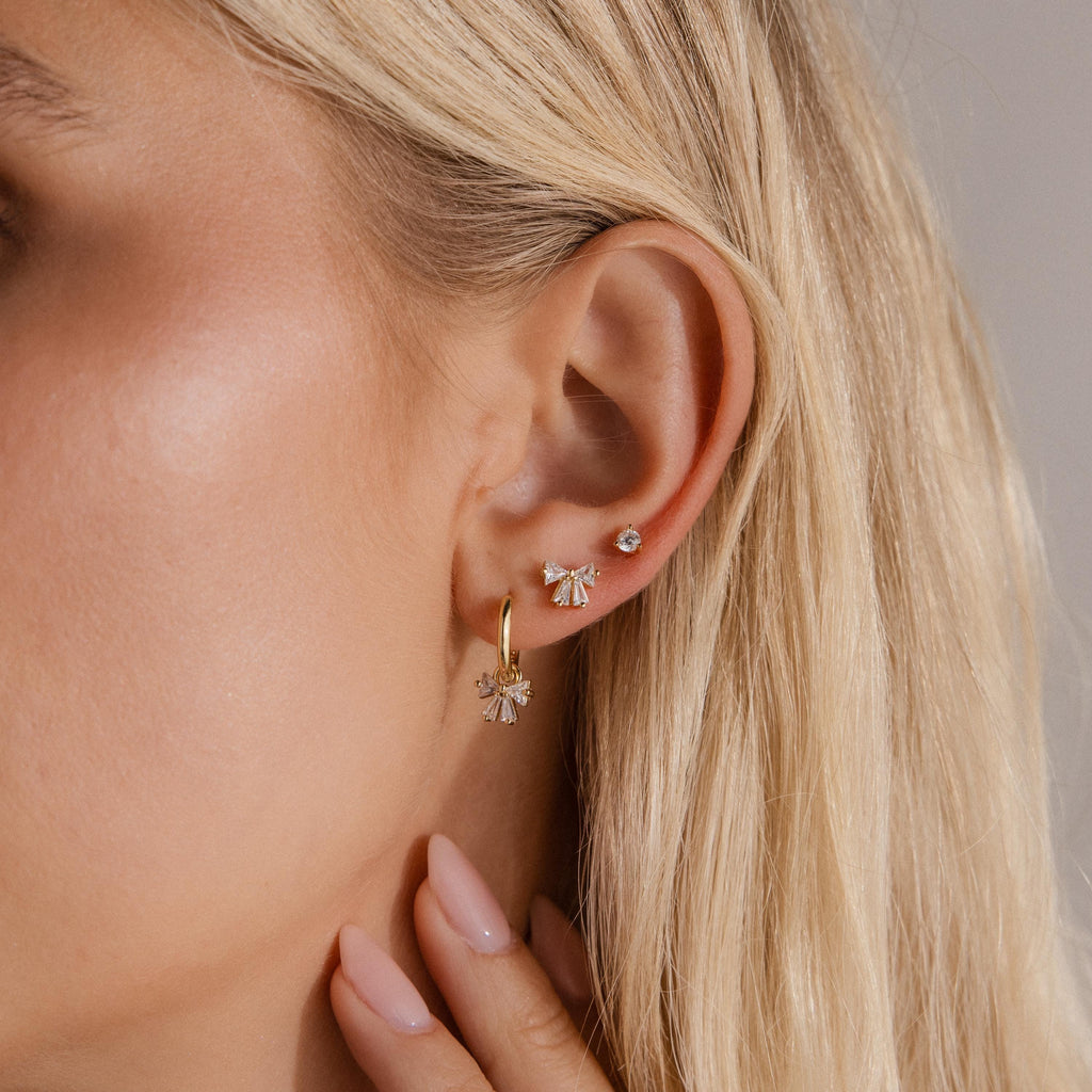 Close up of a woman wearing a gold Starlight Bow Hoop earring paired with a matching Starlight Bow stud and a small crystal stud, styled together for an elegant layered ear look.