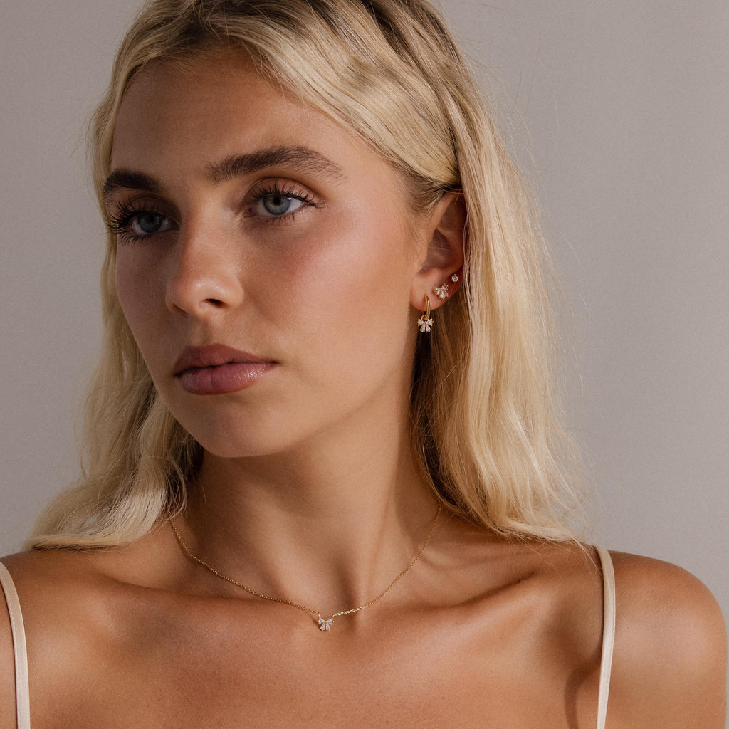 A woman with long blonde hair, wearing gold jewelry and the Diamond Bow Studs with a cream camisole, gazes slightly to the side.