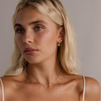 A woman with long blonde hair, wearing gold jewelry and the Diamond Bow Studs with a cream camisole, gazes slightly to the side.