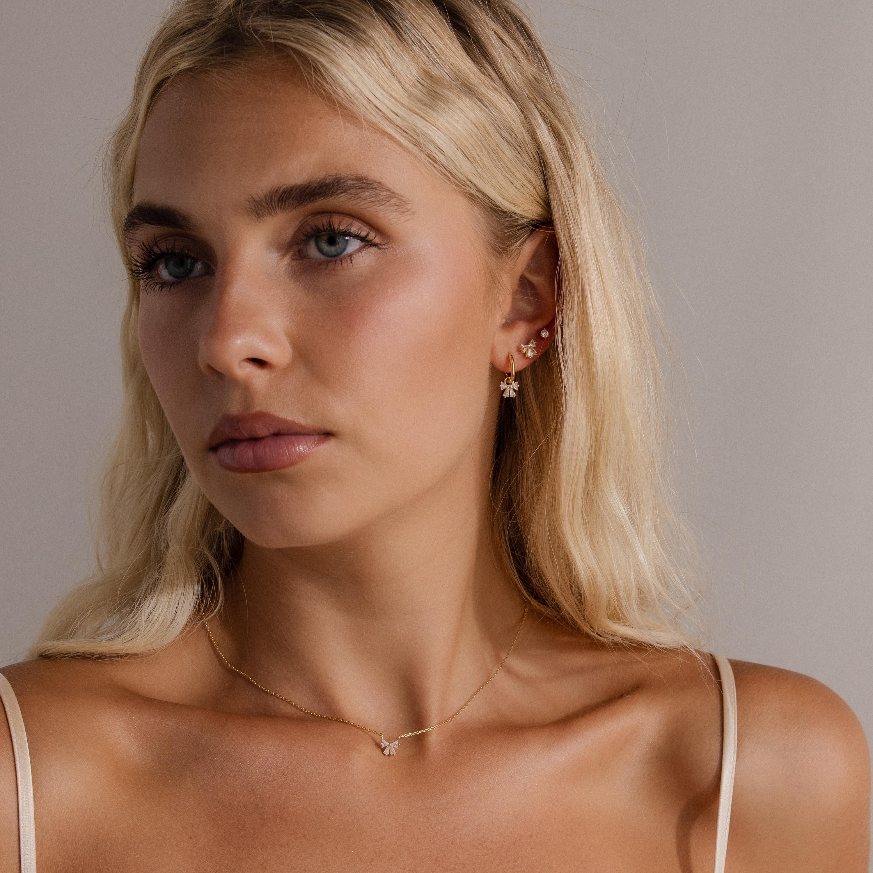A woman with long blonde hair, wearing gold jewelry and the Diamond Bow Studs with a cream camisole, gazes slightly to the side.